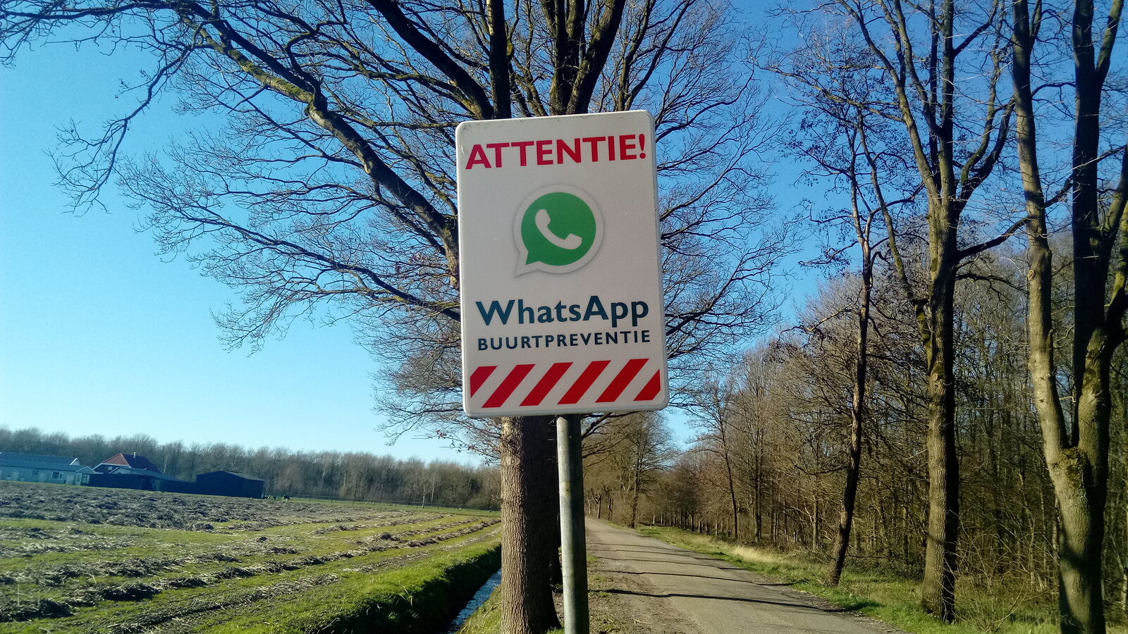 Un cartel de «Attentie! WhatsApp Buurtpreventie» junto a un camino rural en los Países Bajos.